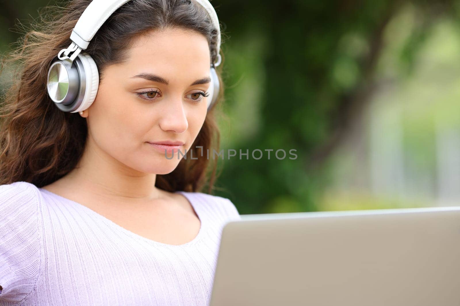 Concentrated woman with wireless headphones watching media on laptop in a park
