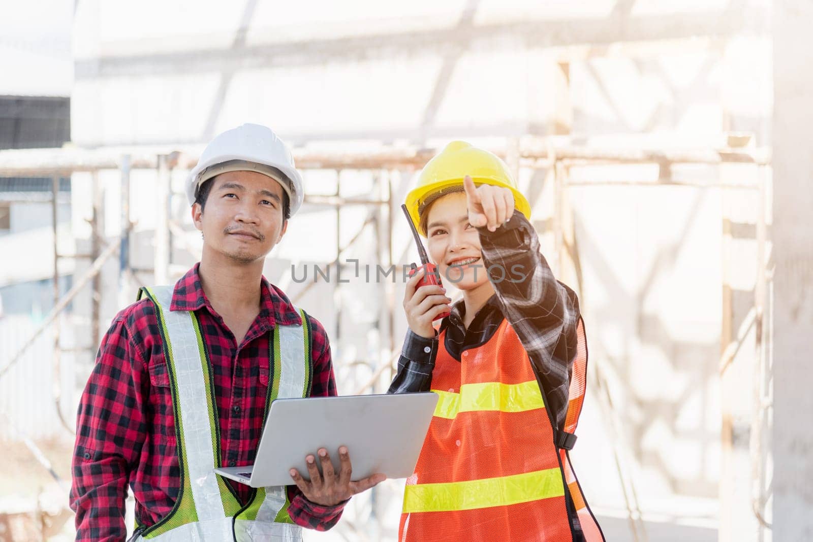 Asian engineer foreman worker man and woman working at building construction site use laptop and talking with radio, engineering hold computer and radio discuss and control worker employee to building