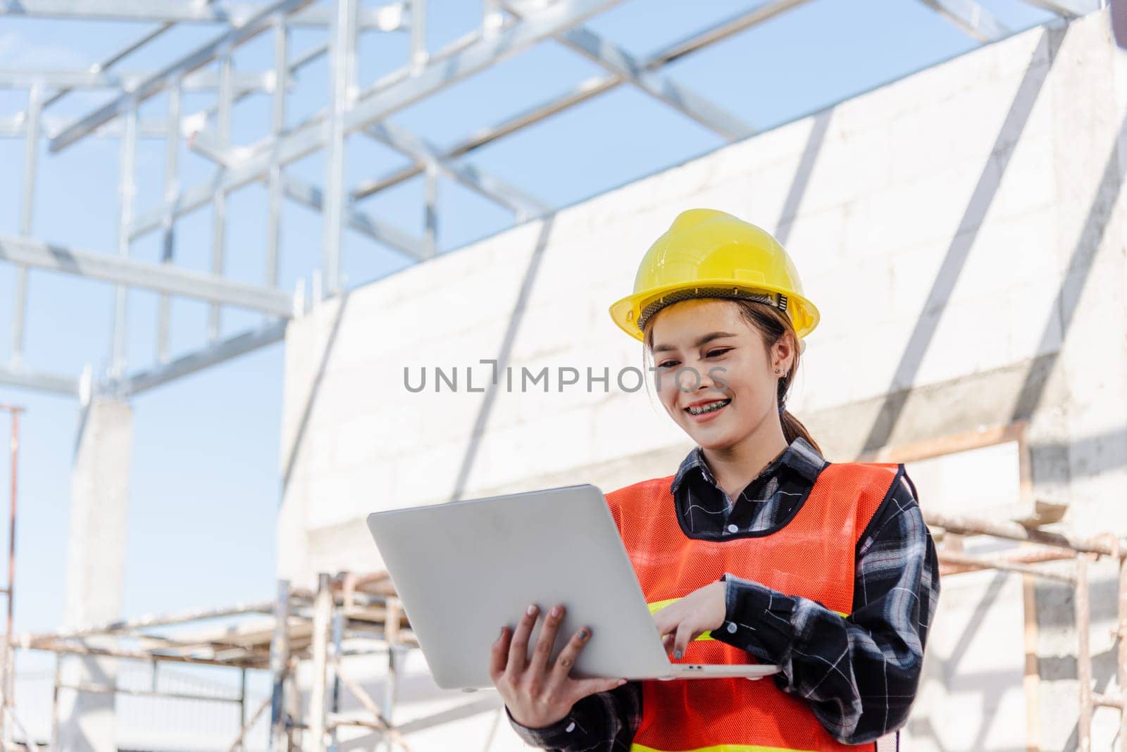Asian engineer architect worker woman holding laptop inspect and oversee infrastructure progress at construction site, engineering use computer to operate project and control worker to building