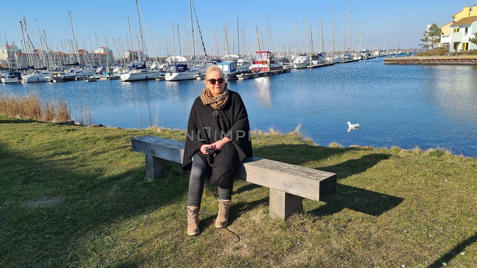 Portrait of a blonde woman wearing sunglasses sitting on a bench in the marina with boats in the background