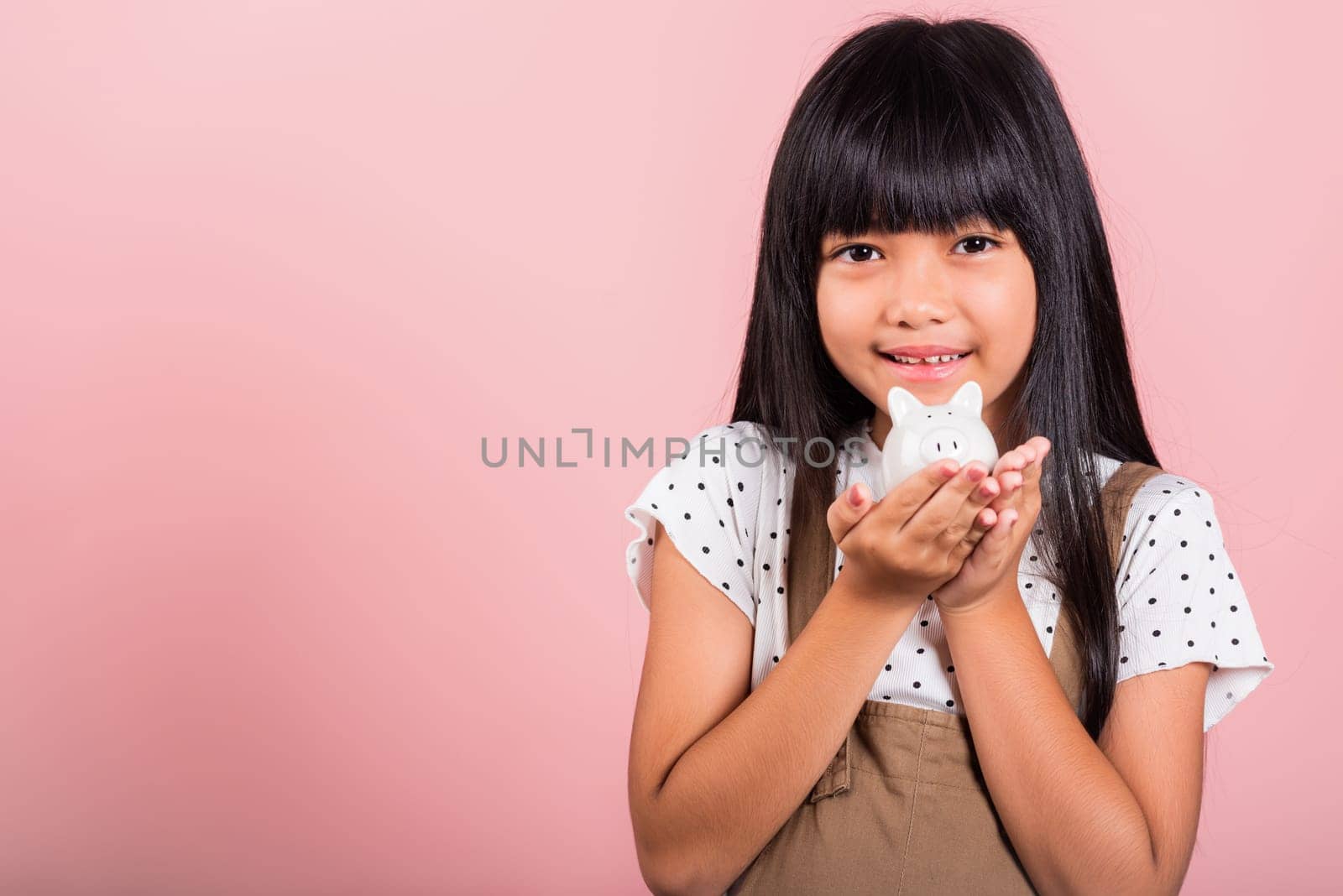 Asian little kid 10 years old holding piggy bank and looking at camera at studio shot isolated on pink background, Happy child girl lifestyle smiling with is full piggybank, Personal money savings