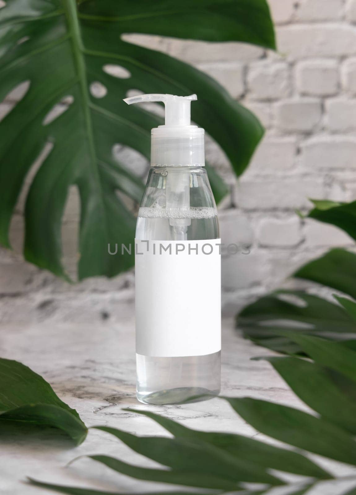 Trasparent one pump dispenser and tropical monstera leaves on marble table close up. Brand packaging mockup. Skincare beauty product. Natural herbal cosmetics concept..