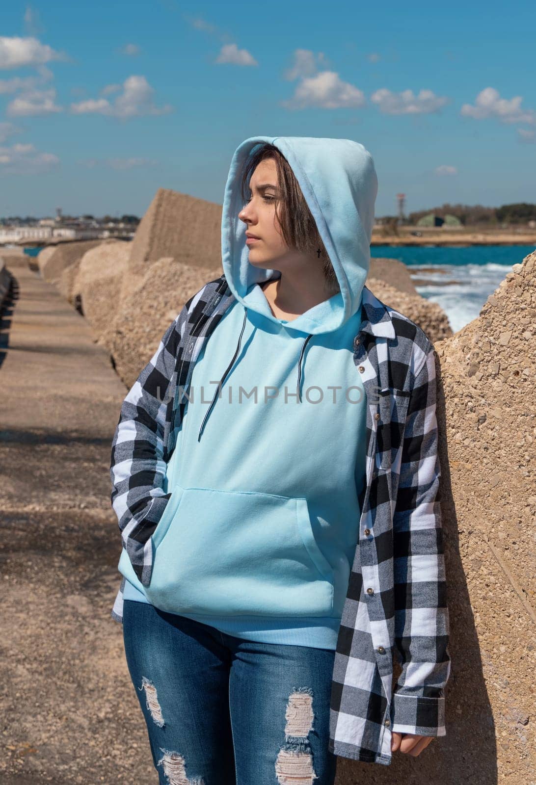 Teenager in light blue hoodie and plaid shirt looking aside. Teen girl stays outdoor near the sea, concrete blocks and blue sky, windy day. Clothing mockup, copy space