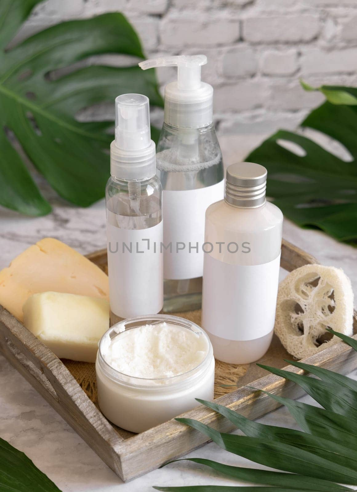 Cosmetic bottles and skin care accessories near green monstera leaves close up against white brick wall. Brand packaging mockup. Organic beauty products. Natural homemade cosmetic ..