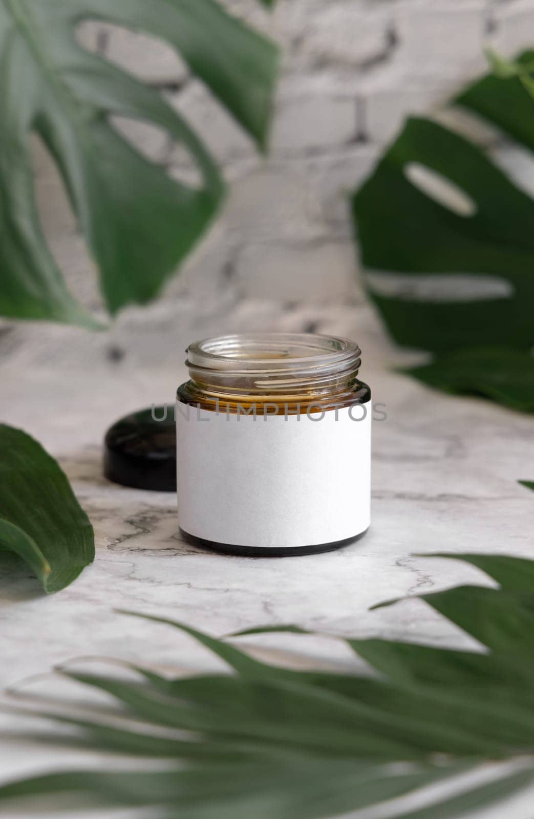 Opened Brown jar and monstera leaves on marble table close up. Brand packaging mockup. Skincare beauty product, cream or butter. Making homemade herbal cosmetics from natural ingredients..