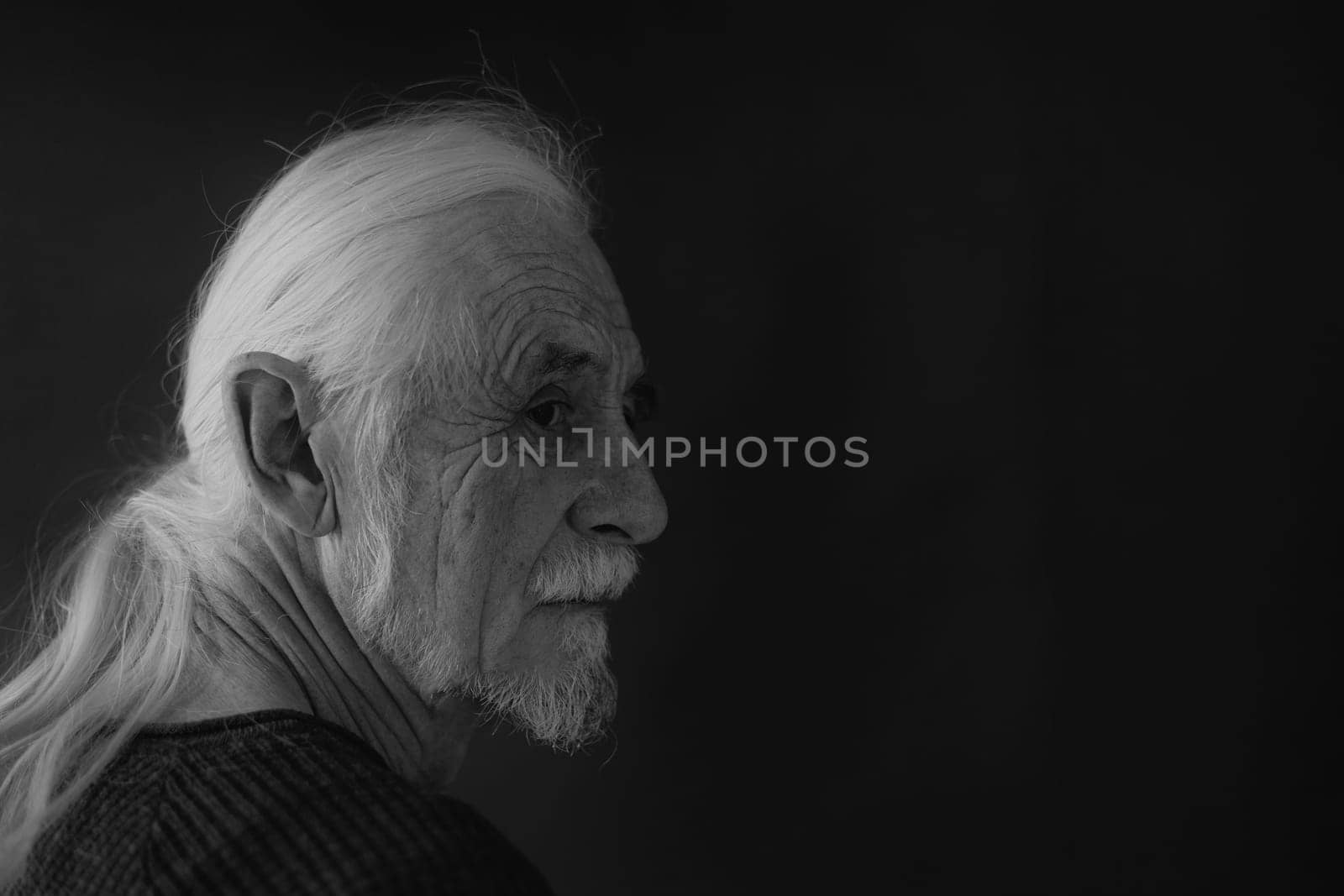 Low key studio black and white portrait of beautiful gray hair old man looking over his shoulder at the camera. Horizontally.
