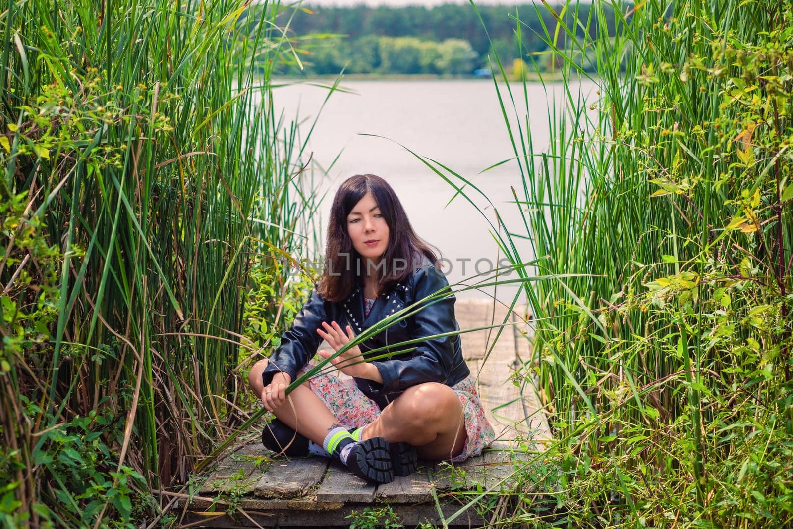 A black-haired young girl in a black jacket sits on a wooden bridge on the lake shore