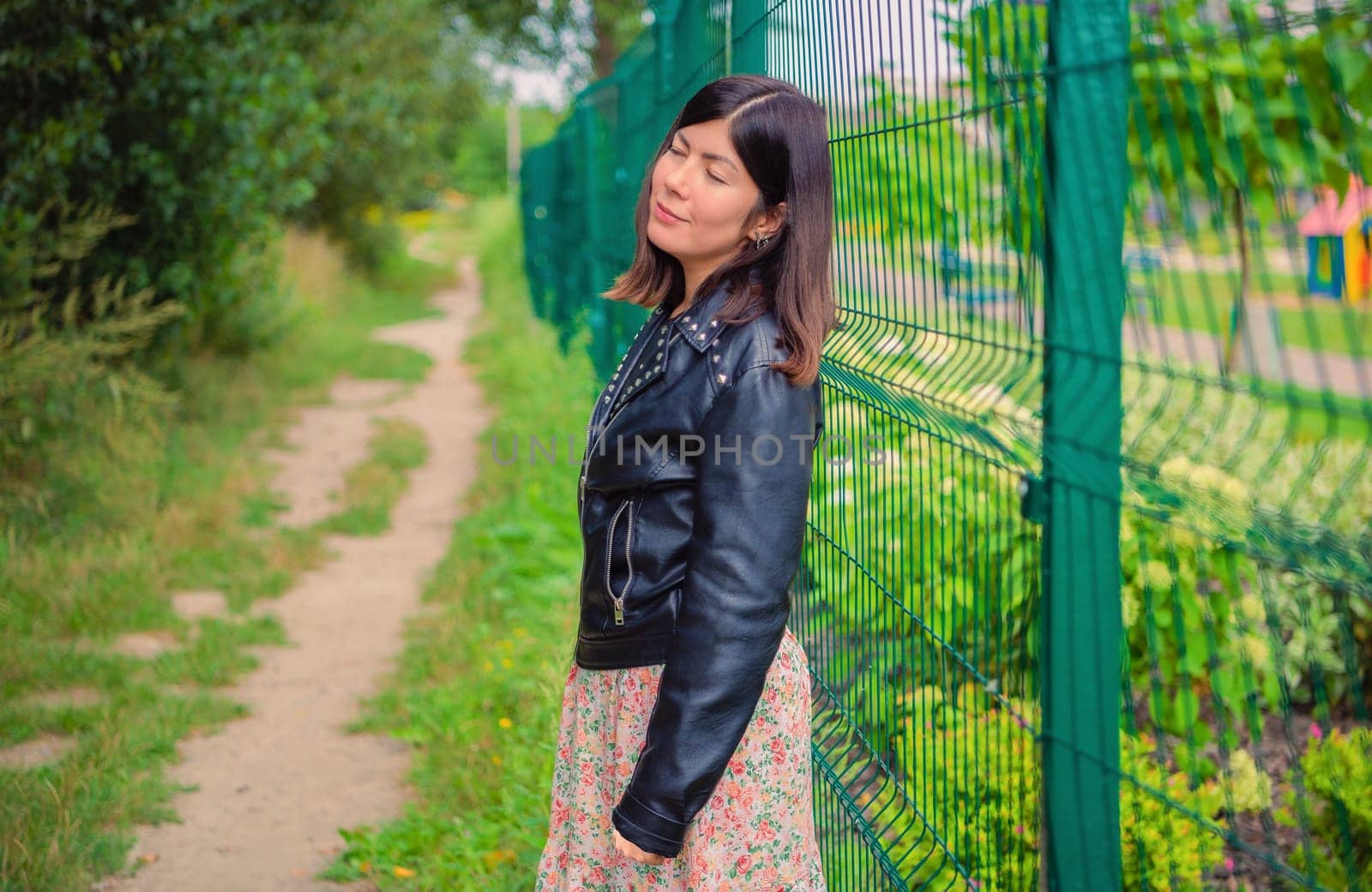 A cute girl in a black jacket stands with her eyes closed, leaning her back against a green iron fence.