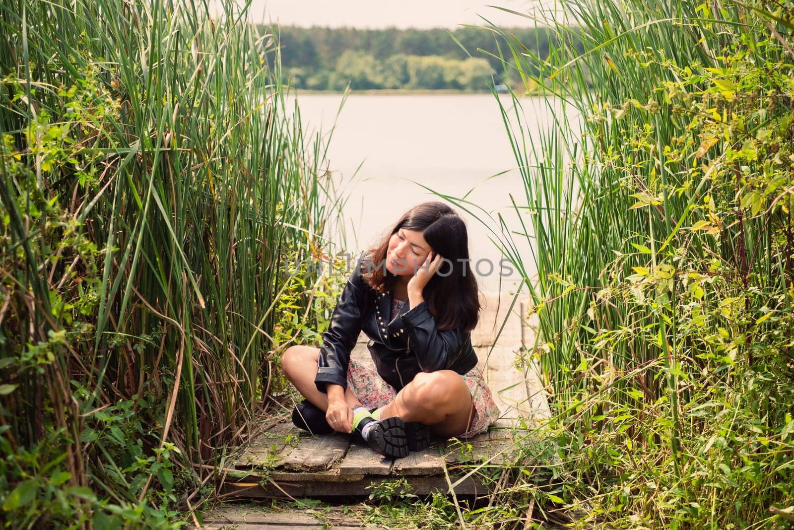 a brunette girl in a black jacket sits on a wooden bridge on the bank of a pond, propping her head up with her hand