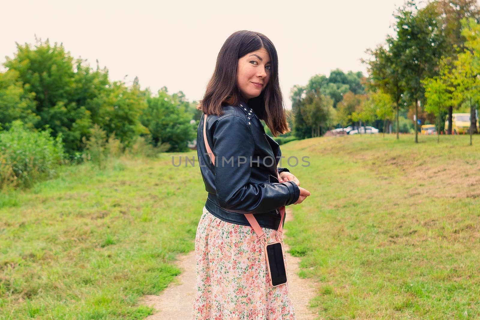 beautiful smiling girl with a phone on her belt looks back while standing in the middle of a green park