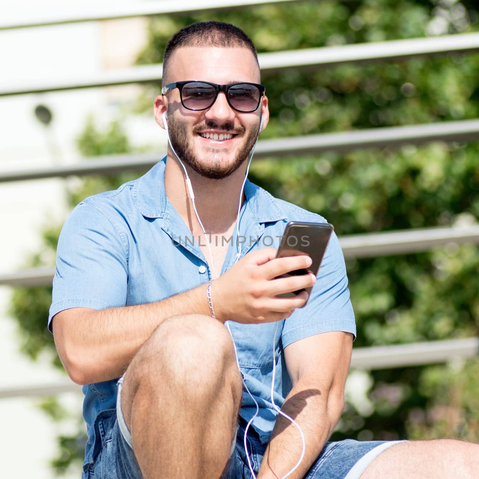 The smiling young guy with earphones is using phone for listening music outdoors.