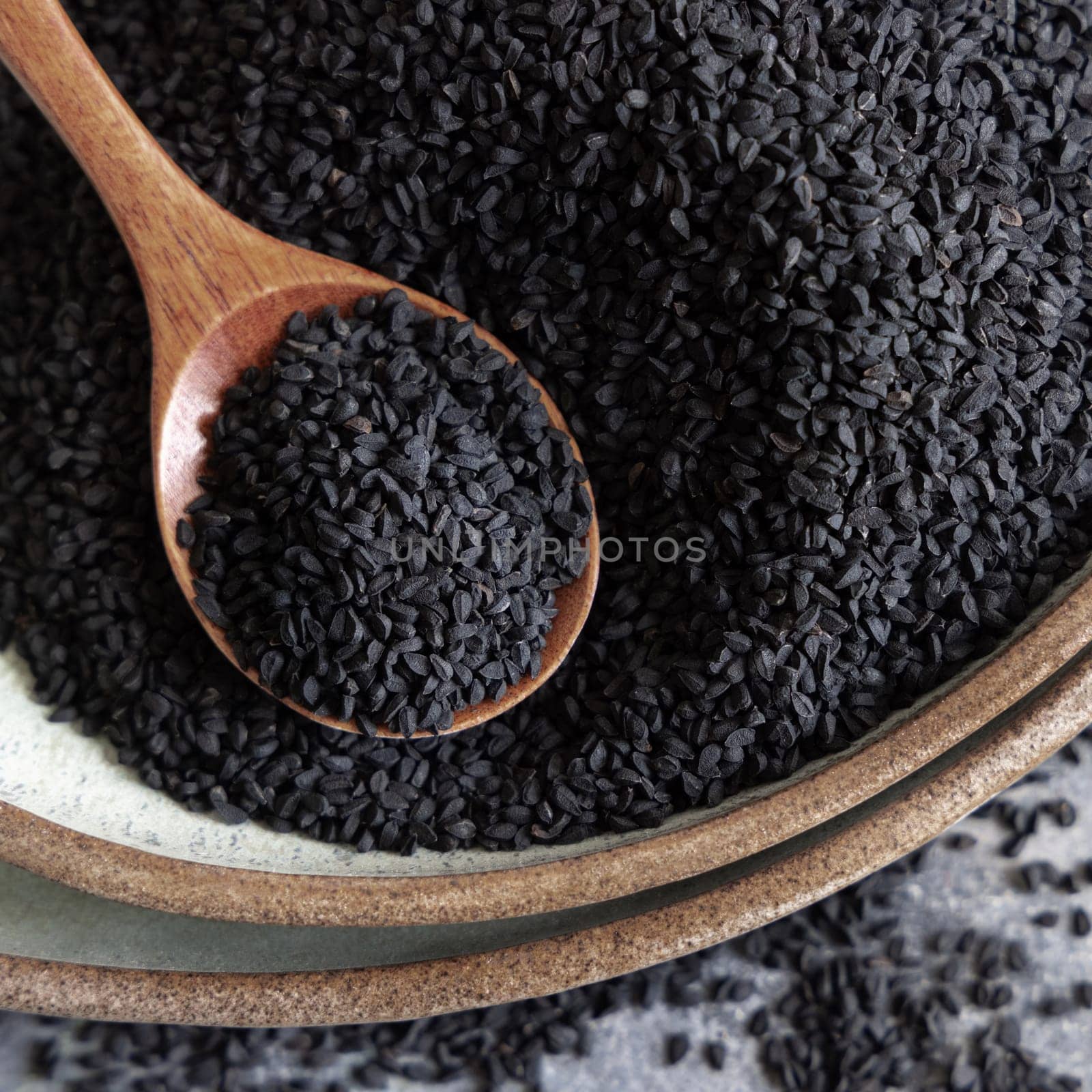 Plate of Indian spice Black cumin (nigella sativa or kalonji) seeds on grey table with a wooden spoon, top view. Traditional asian medicine, healthy and vegetarian food concept
