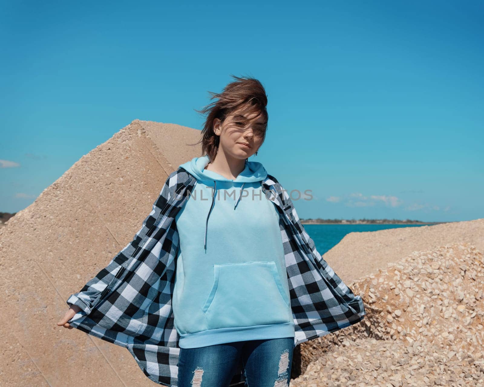 Short haired teenager girl in light blue hoodie and plaid shirt looking into the camera. Teen girl stays outdoor against concrete blocks and blue sky, windy day. Clothing mockup, copy space