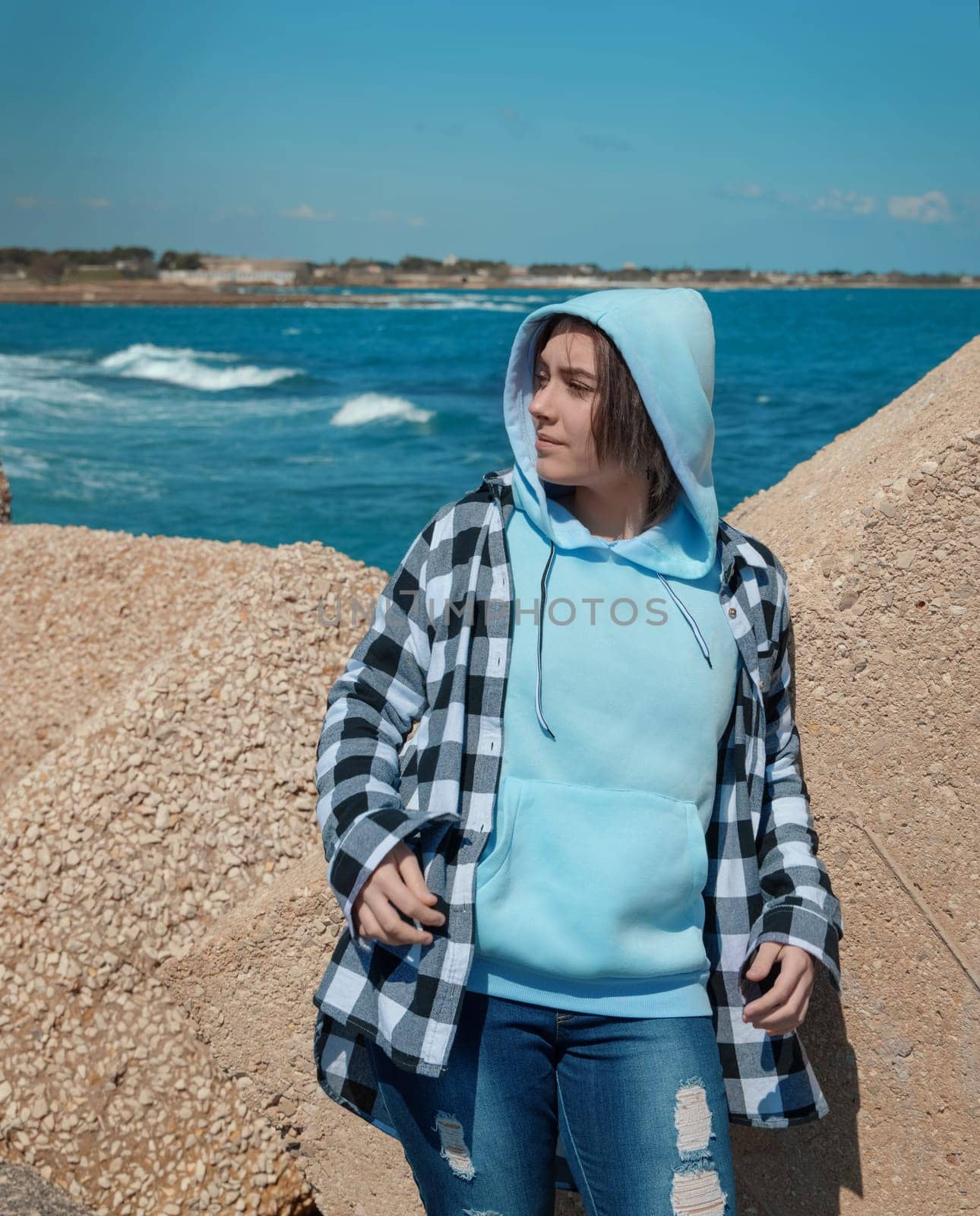 Teenager in light blue hoodie and plaid shirt looking aside. Teen girl stays outdoor near the sea, concrete blocks and blue sky, windy day. Clothing mockup, copy space