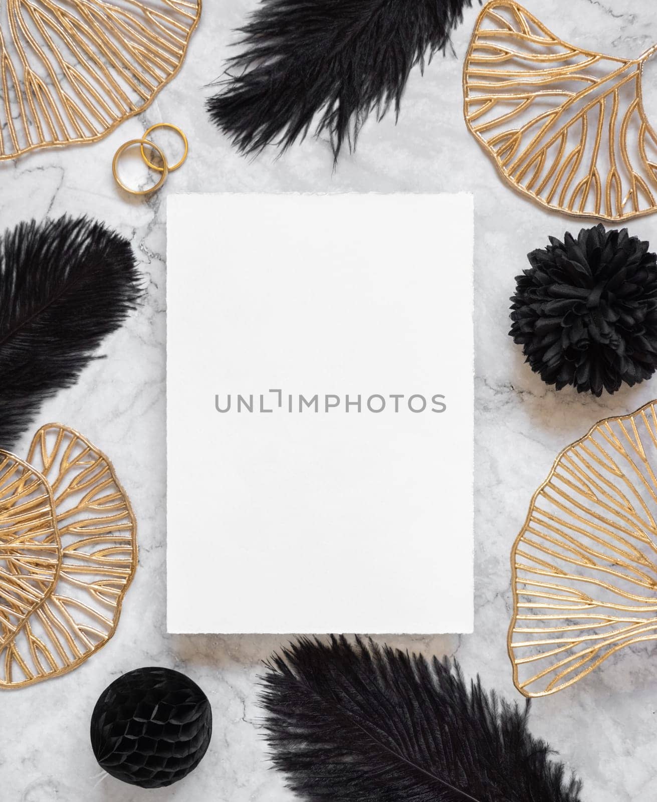 Blank card on marble table near black feathers and golden leaves with wedding rings top view. Greeting or Invitation card mockup. Elegant femminine flat lay with vertical paper card
