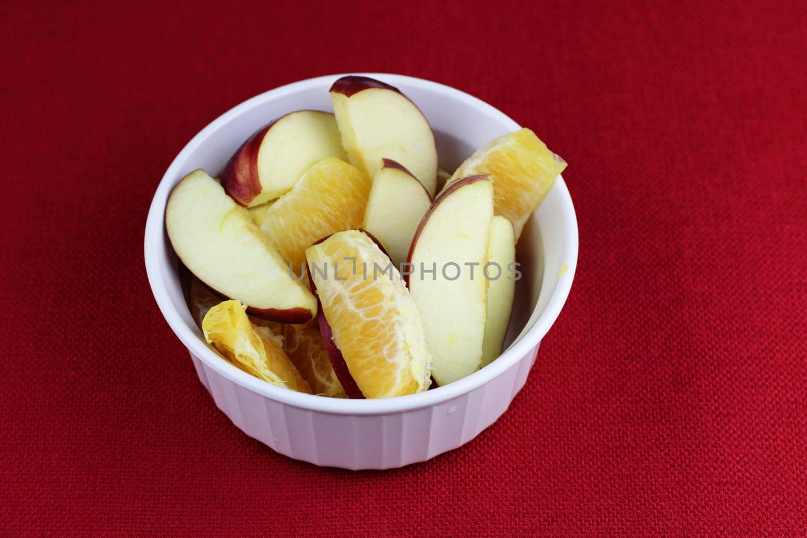 White breakfast fruit of red apple and orange sections on a red place mat close-up. Breakfast food fruit slices of orange citrus and red apple food in a round white bowl on a red background placemat.
