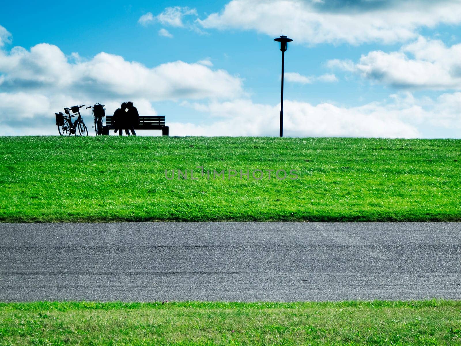 Landscape scene with a pair of lovers as silhouettes against the light on a park bench on the dyke with bicycles and a lantern next to them.