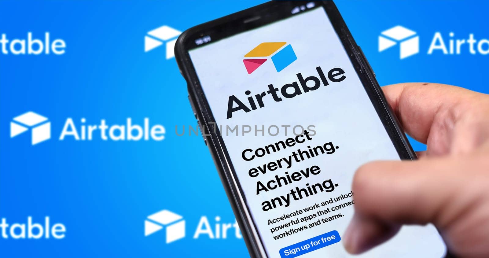 San Francisco, CA, US, May 2022: Male hand holding a phone with Airtable company web page on screen. In the background the logo Airtable blurred and repeated. Airtable is a cloud collaboration service