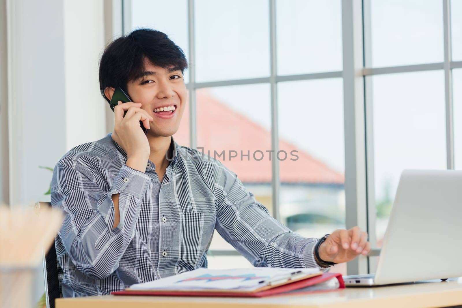 Asian businessman smiling and talking on a mobile phone at home office desk. The man smile call on smartphone with customer at the house, making sell offer
