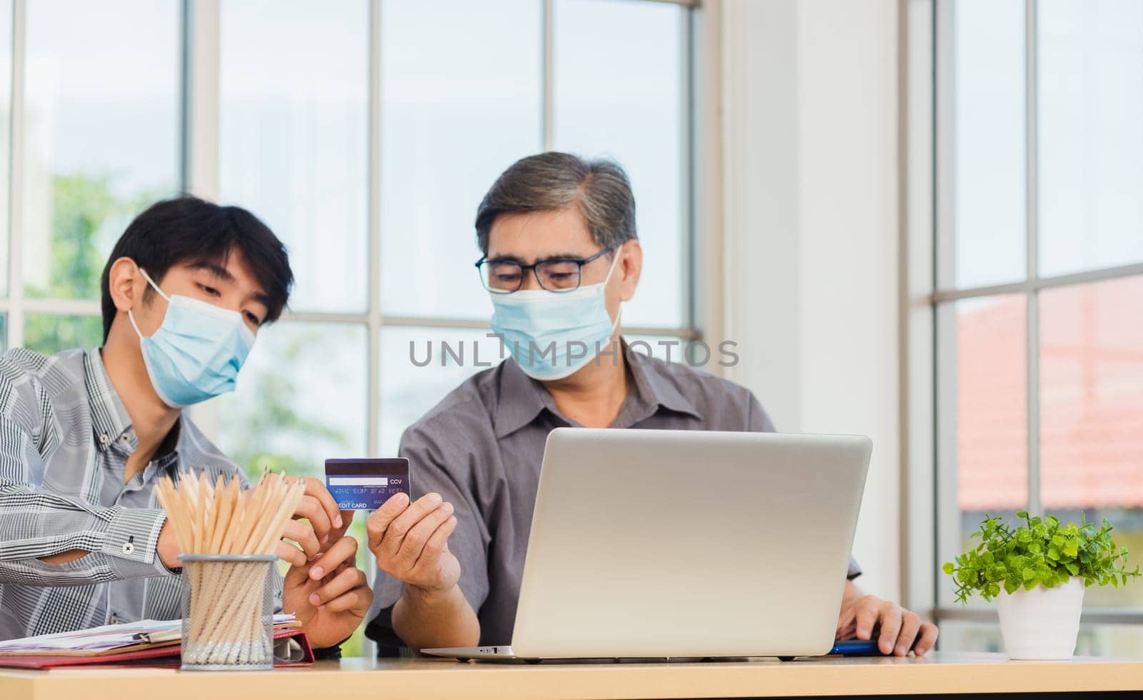 Asian young man teaching senior father how to use credit card for shopping online with laptop computer in the living room, adult son holding bank card helping Dad to purchases web online application