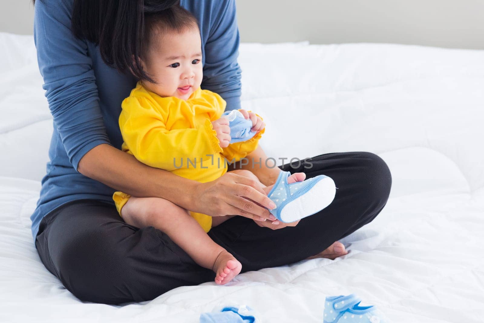 Asian young mother sitting on the bed wearing shoes to his cute baby little girl toddler on bedroom, Mom helping little infant wears shoes