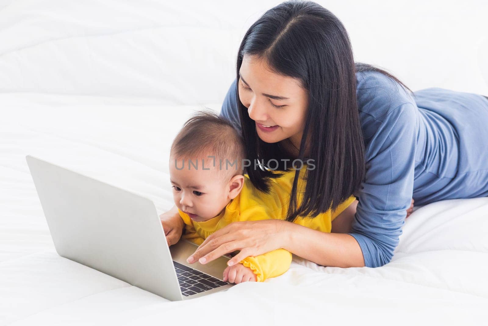 Asian happy young mother and baby lifestyle working on laptop, woman mom and daughter reading and learning technology on screen while notebook computer on bedroom white bed