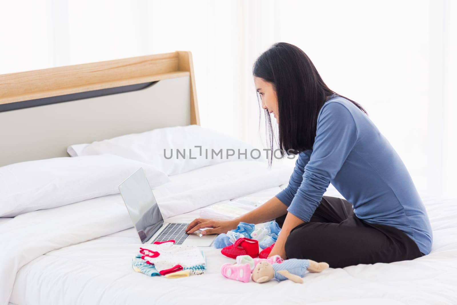 Asian mother shopping online for her baby clothes on laptop computer and tablet on the bed she makes purchase new baby clothes for an unborn baby preparing for the new child