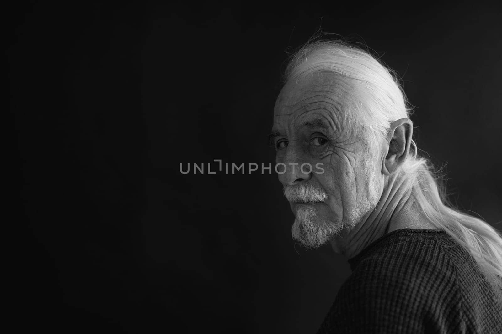 Low key studio black and white portrait of beautiful old man with gray hair in a braid looking over his shoulder at the camera. Horizontally.
