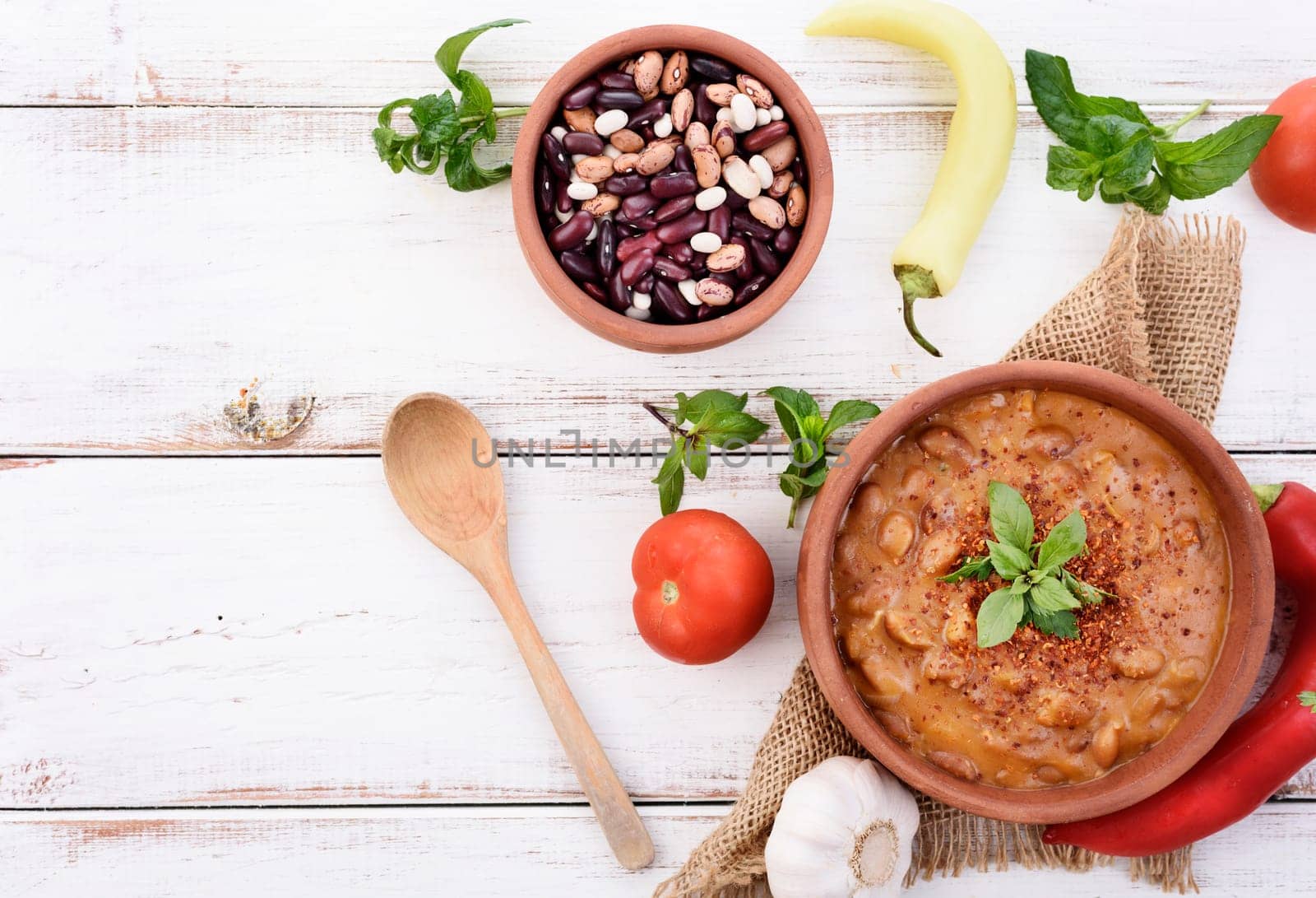 Traditional homemade recipe for spicy baked beans cooked in vintage clay bowl with vegetable and herbs. Top view of prepared dish and ingredients on white wooden table with copyspace