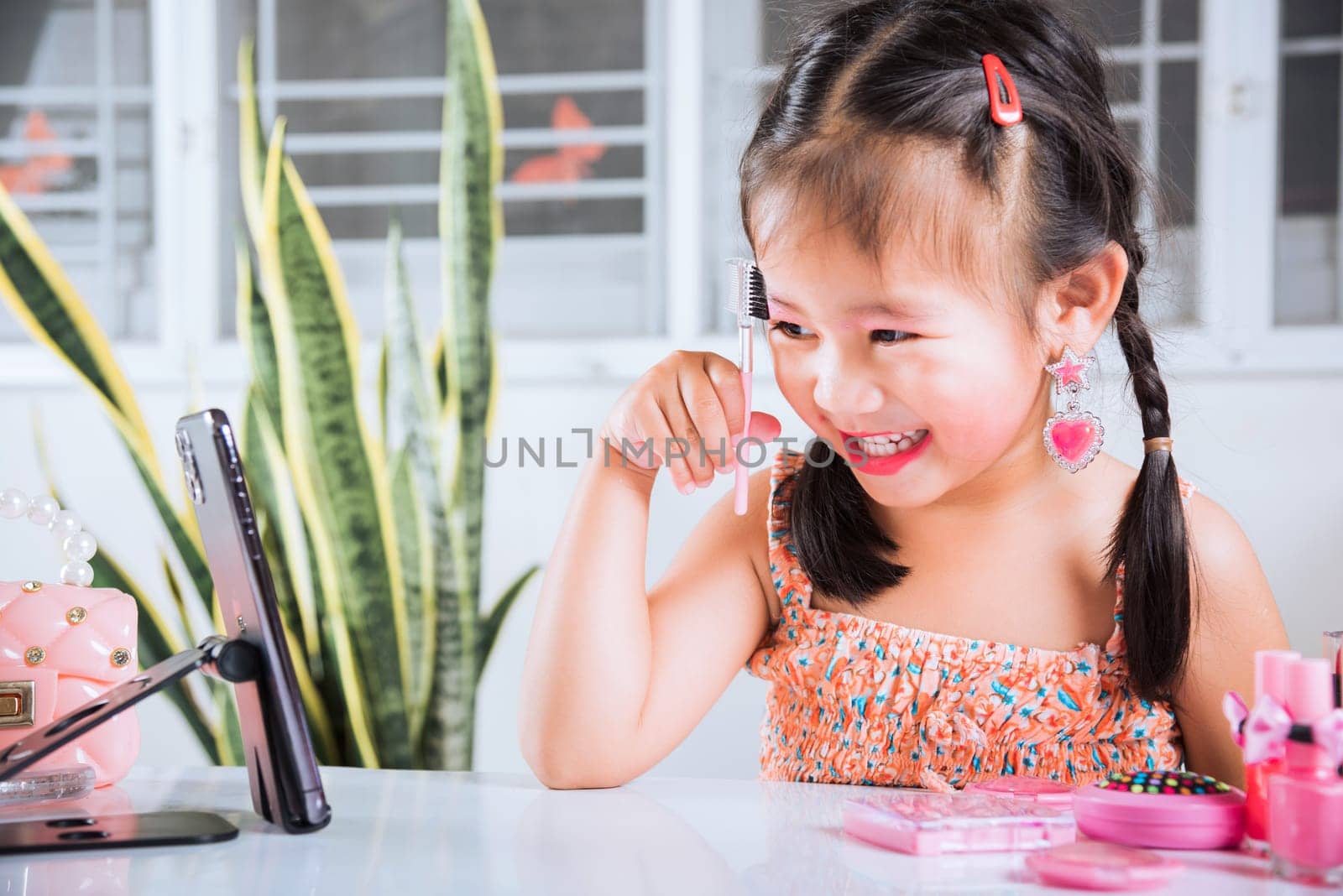 Asian funny little girl making makeup her face she look in smartphone and use eyebrow mascara, Learning activity to be woman, social media blogger recording vlog with cosmetics, online tutorial