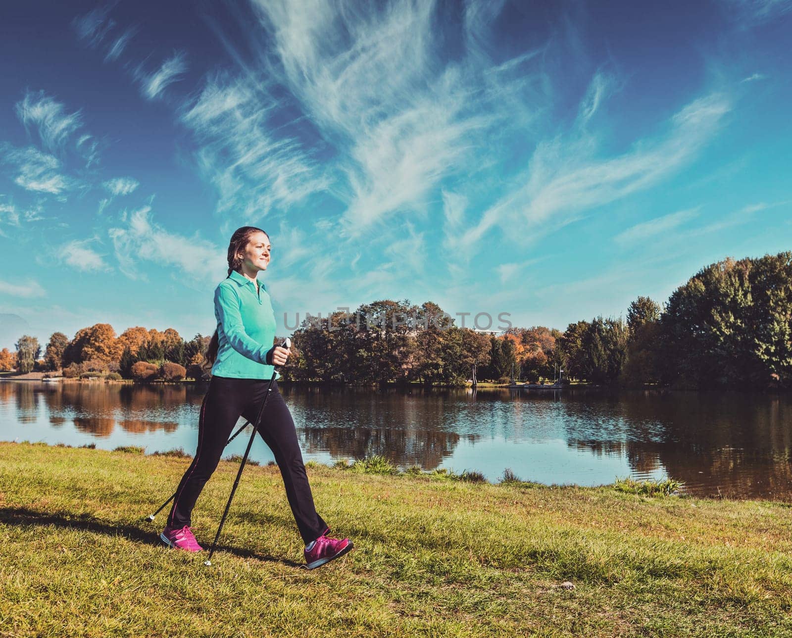 Vintage retro effect filtered hipster style image of nordic walking adventure and exercising - young woman hiking with nordic walking poles in park along river