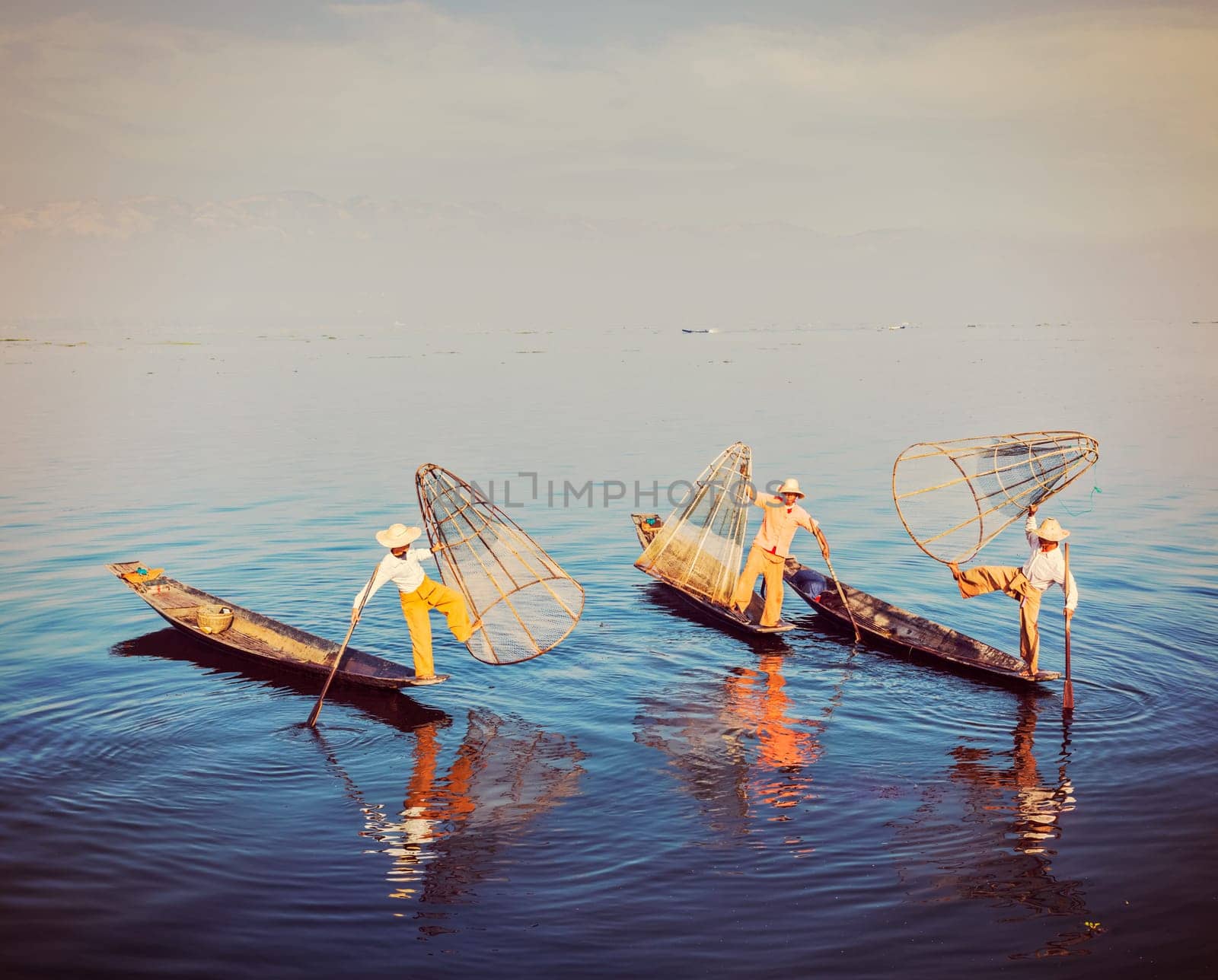 Myanmar travel attraction - Traditional Burmese fishermen balancing with their fishing net on boats at Inle lake in Myanmar famous for their distinctive one legged rowing style. Vintage filtered retro effect hipster style image