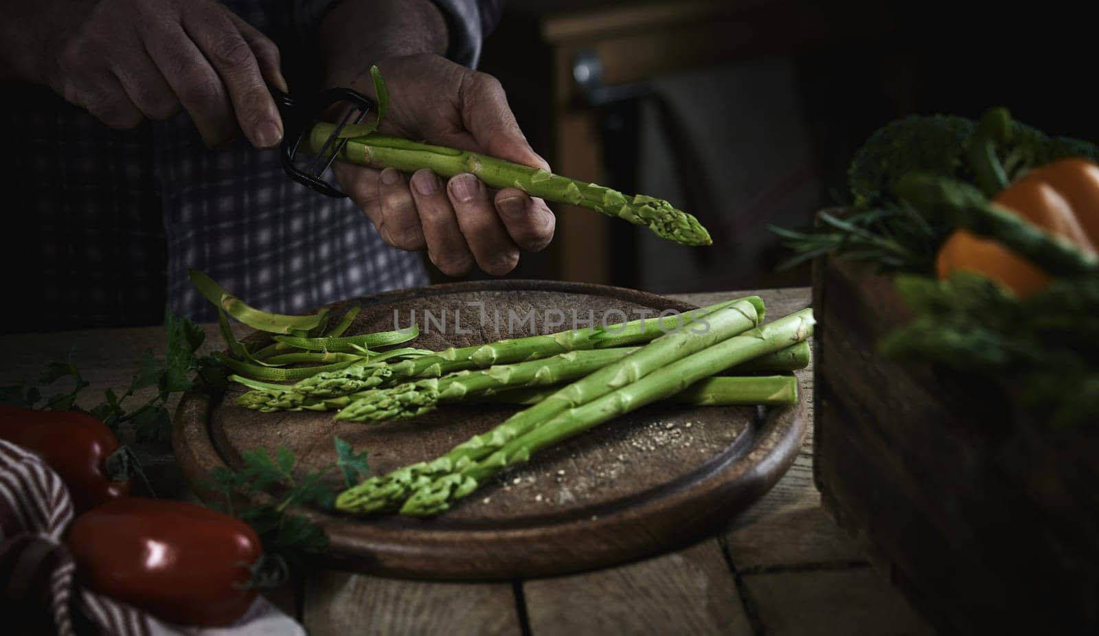 High angle of crop unrecognizable male chef peeling fresh ripe asparagus standing at wooden table with assorted vegetables in kitchen