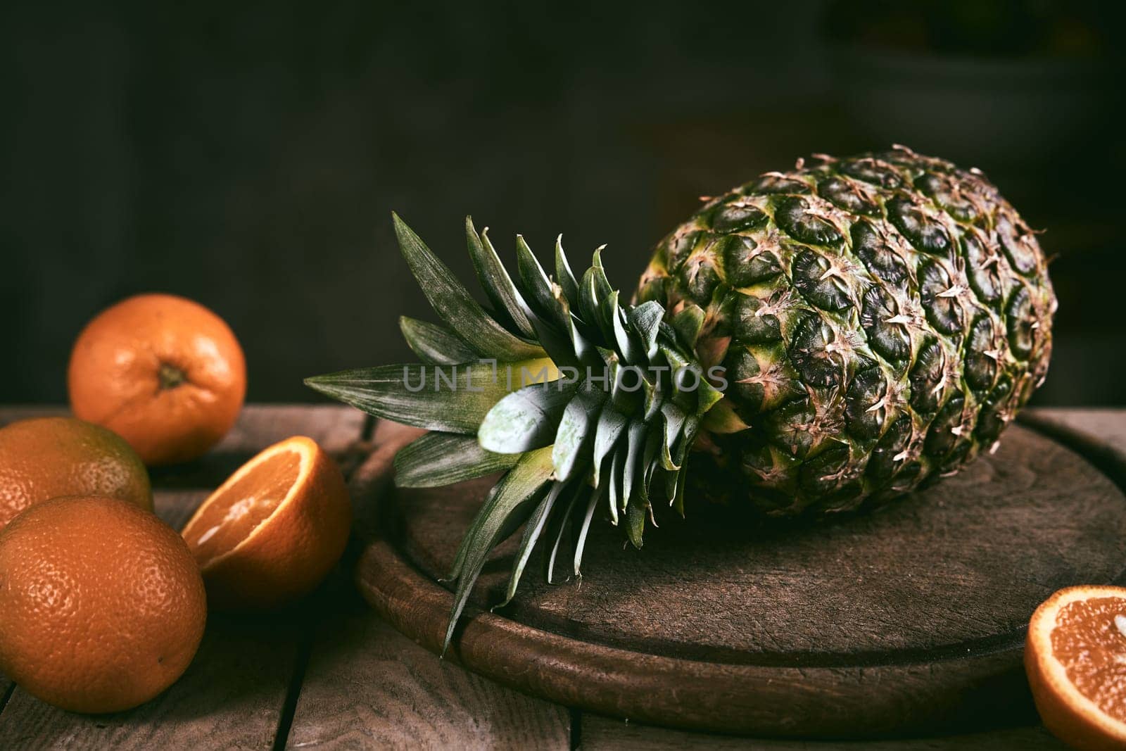Whole ripe pineapple and oranges placed on lumber table near round cutting board against black background in kitchen