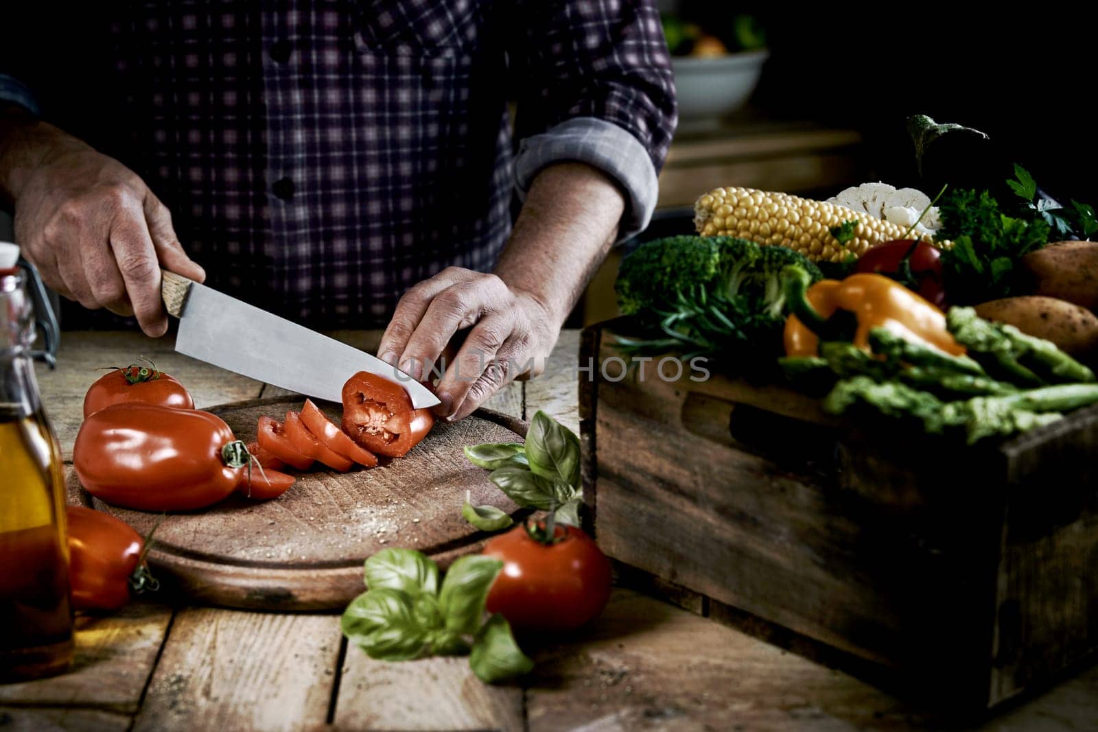 From above of crop anonymous male chef in checkered shirt slicing fresh ripe tomatoes on round shaped wooden cutting board at table with box of assorted raw vegetables in kitchen