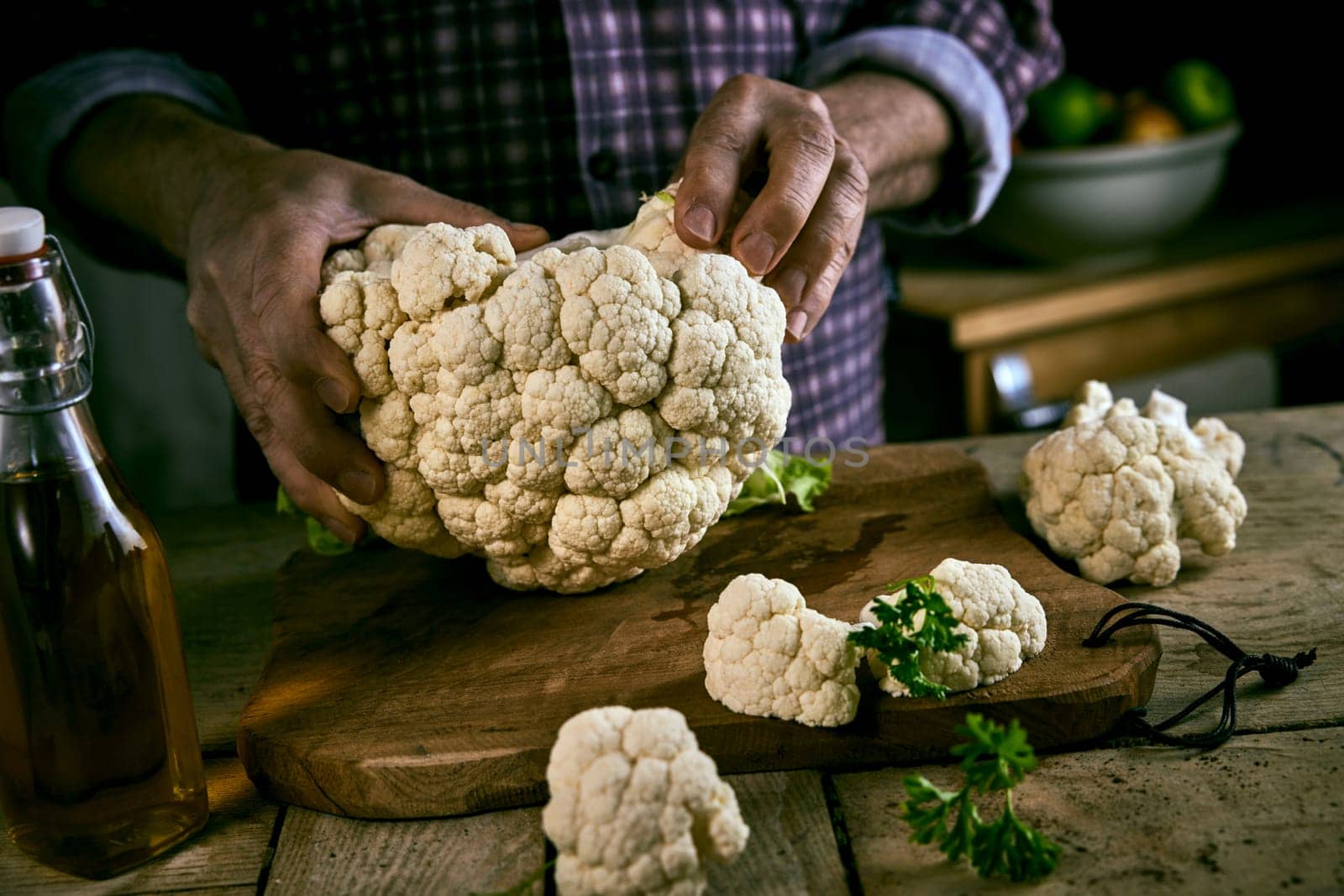 Anonymous man in checkered shirt breaking ripe cauliflower in pieces near bottle of oil while cooking lunch on rustic wooden table