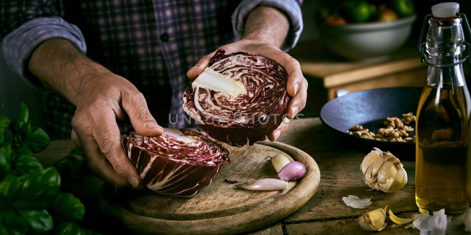 High angle of crop man in checkered shirt putting halves of red cabbage on cutting board near garlic and basil with oil and walnuts during healthy lunch preparation in rustic kitchen