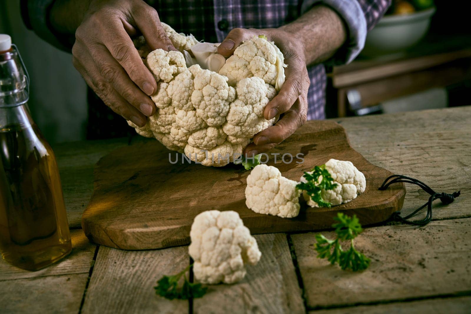 Crop man in checkered shirt tearing fresh cauliflower in pieces over shabby table with wooden cutting board and bottle of oil in rustic kitchen
