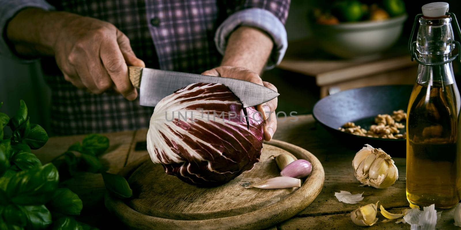 High angle of anonymous male in checkered shirt cutting fresh red cabbage in half with sharp knife near various ingredients while cooking healthy lunch in rustic kitchen at home