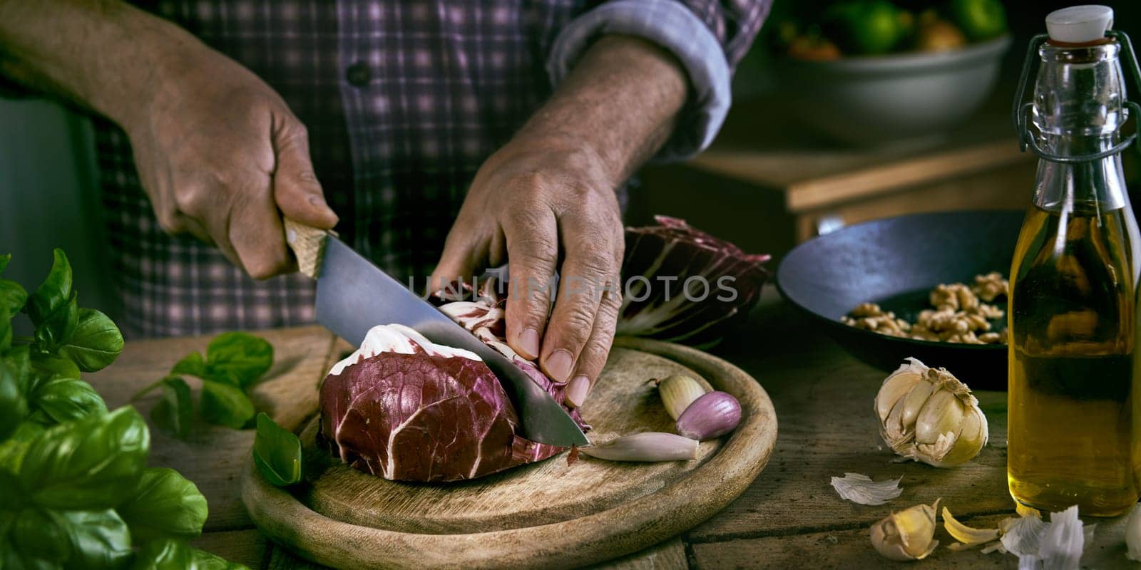 High angle of unrecognizable man in checkered shirt chopping fresh red cabbage on round cutting board near various ingredients during lunch preparation in rustic kitchen