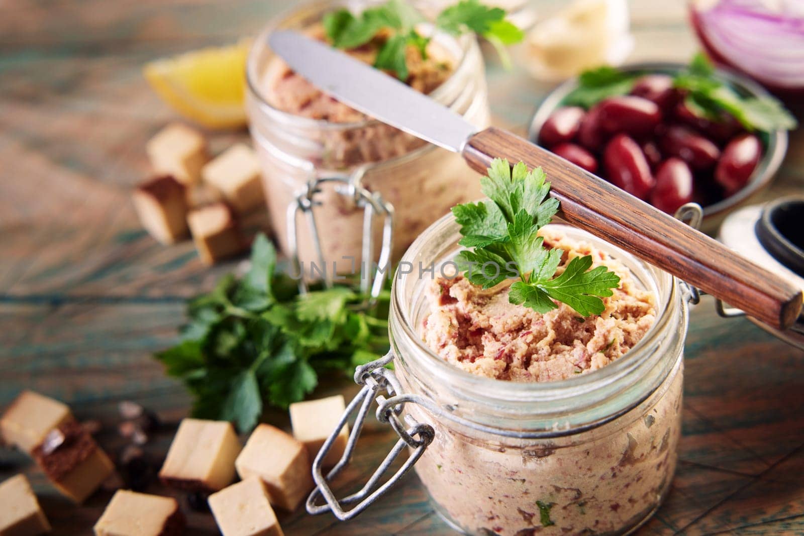 From above of yummy vegetarian liverwurst pate in glass jars decorated with leaves of fresh parsley served on wooden table near bowl of red beans and cubed cheese in kitchen