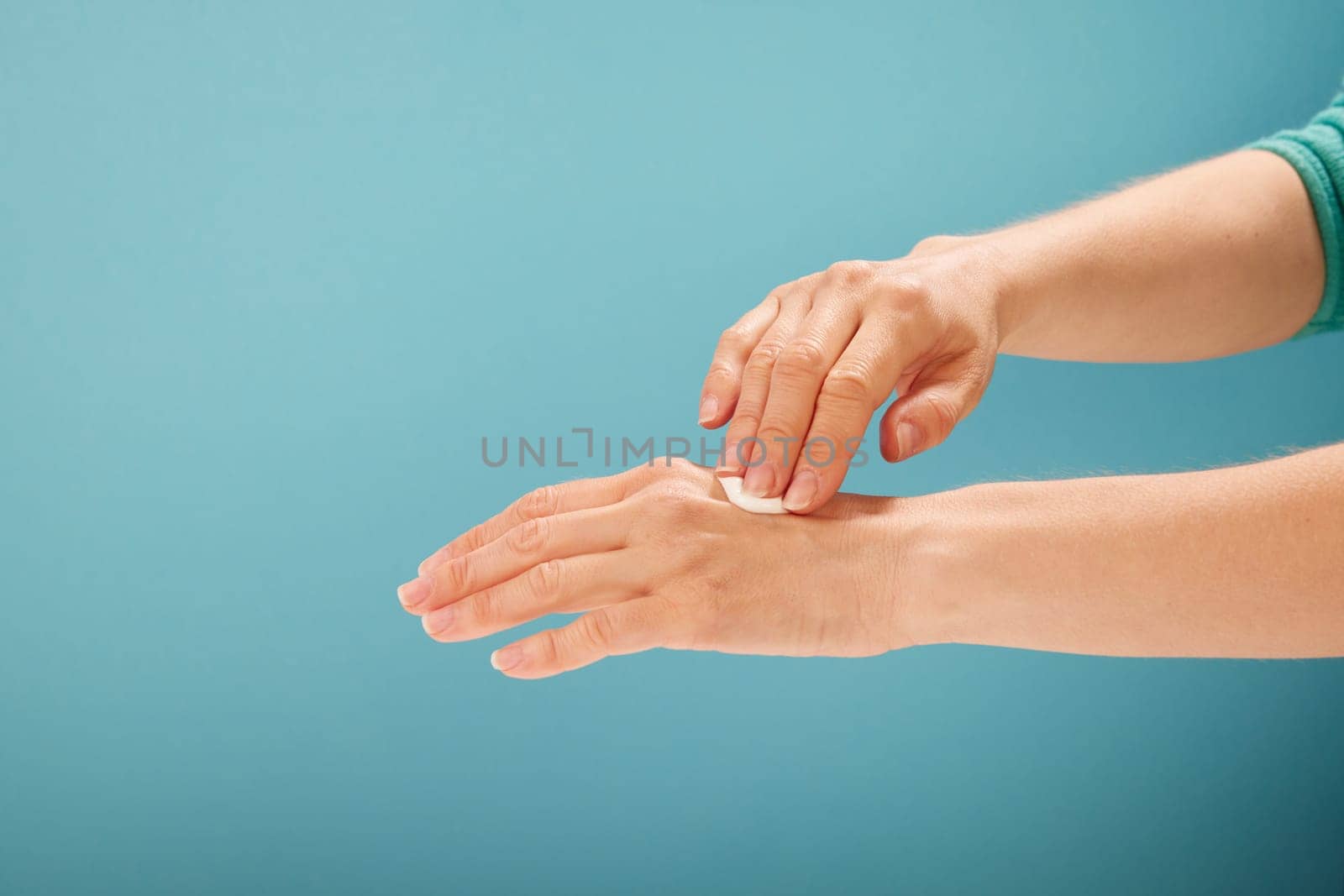 Crop female applying moisturizing cream on hands during spa session against turquoise background