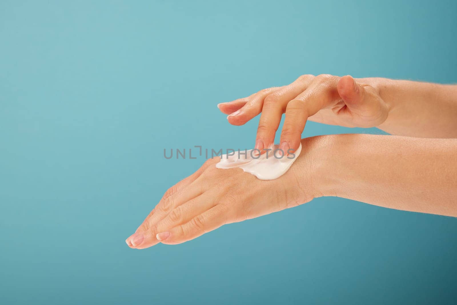 Anonymous woman spreading moisturizing cream on back of hand during skin care routine against turquoise background