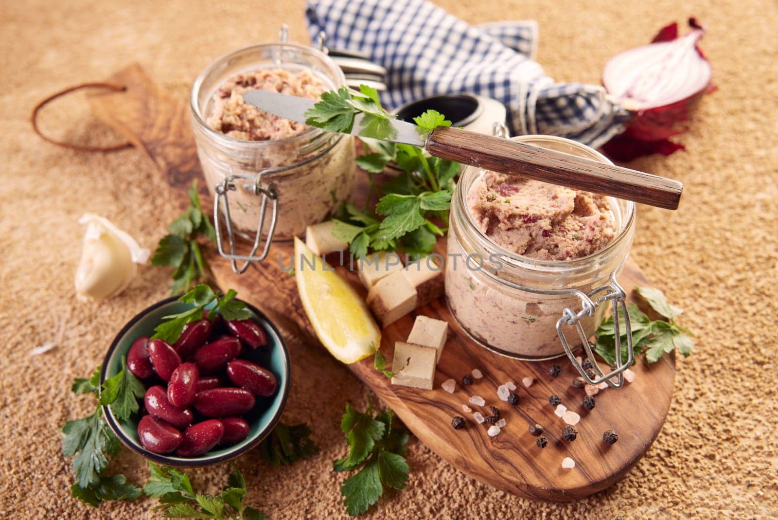 High angle of tasty vegan liverwurst pate in glass jars with cubed cheese lemon and parsley served on wooden cutting board near bowl of kidney beans onion and garlic