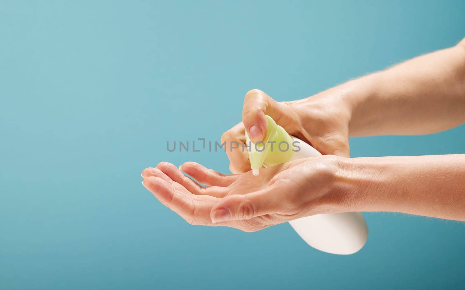 Unrecognizable female squeezing soap from bottle with dispenser on hand during hygienic procedure against turquoise background