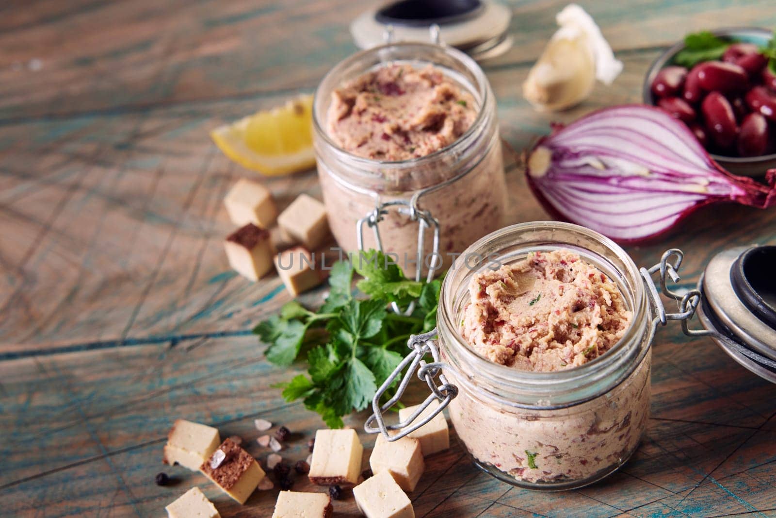 High angle of glass jars with appetizing homemade pate placed on wooden table near scattered cubed cheese herbs and halved onion in kitchen in daylight