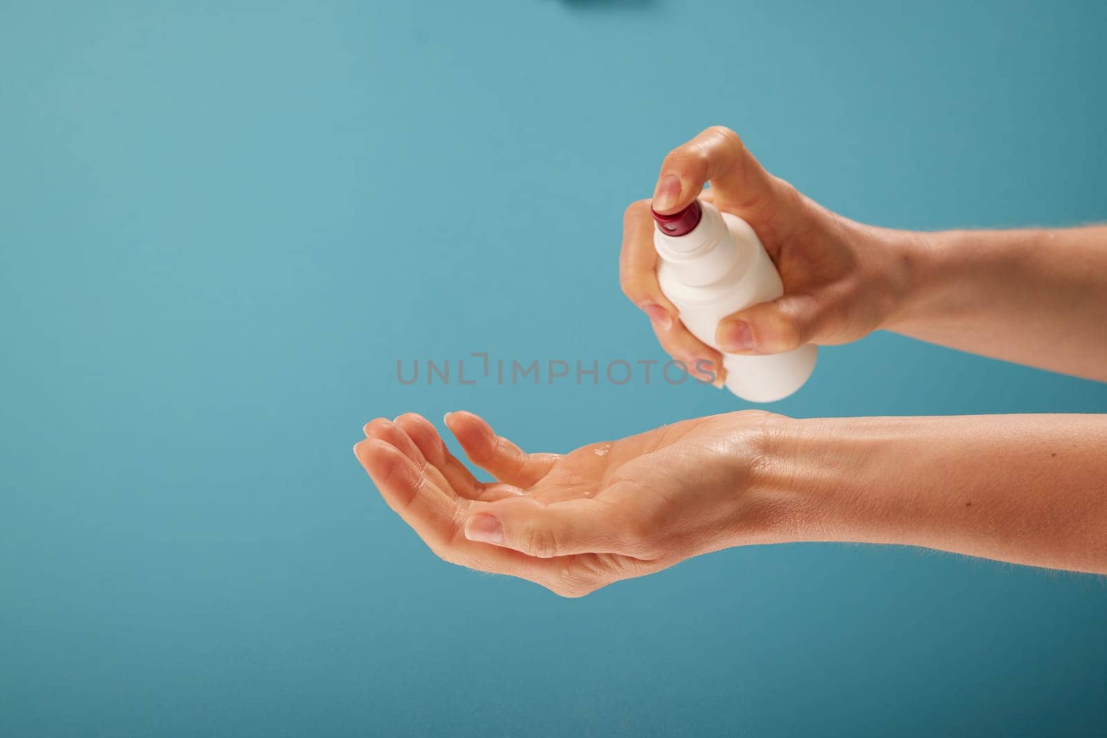 Unrecognizable female disinfecting hands with sanitizer from plastic spray bottle against turquoise background