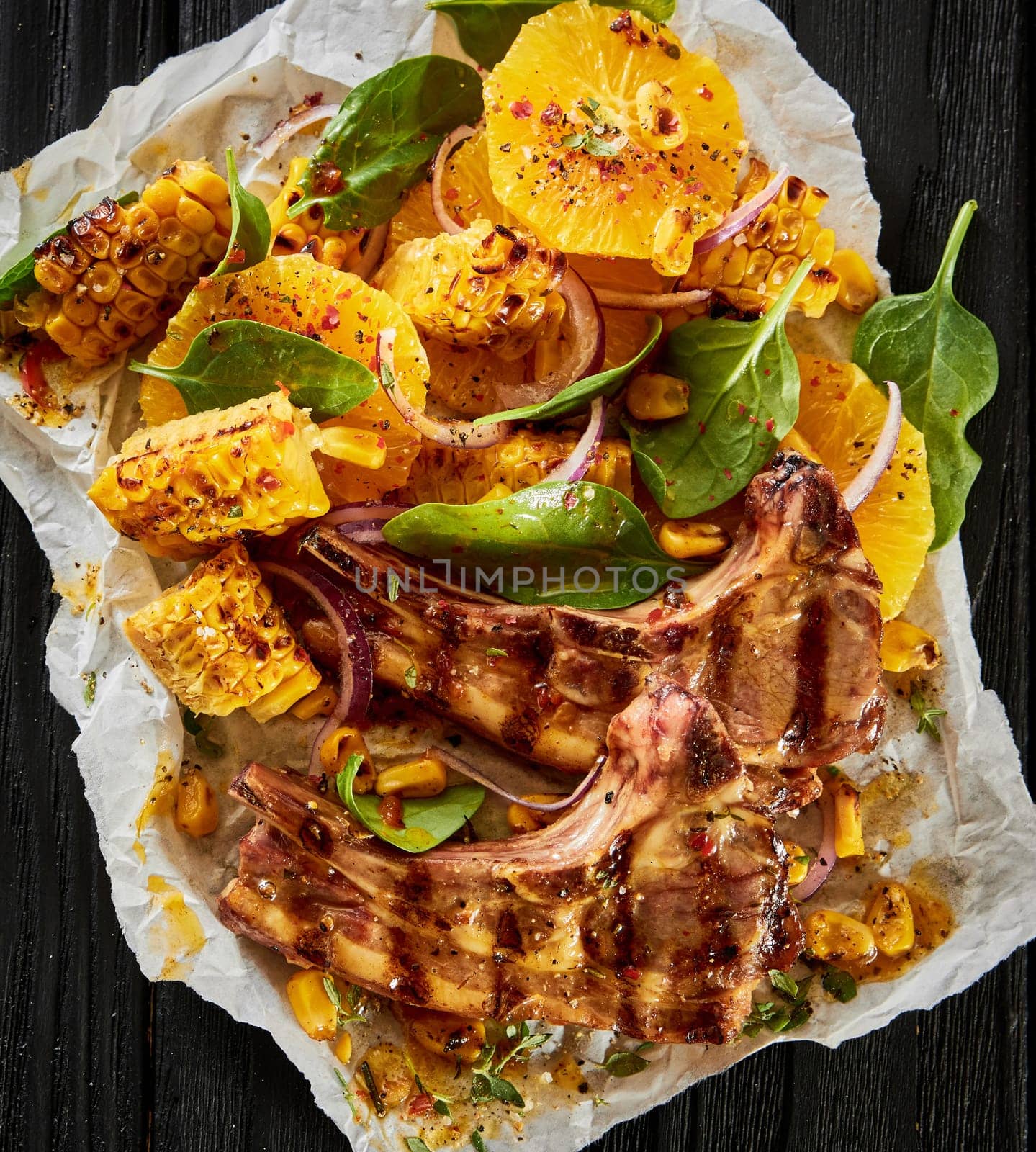 Top view of palatable grilled lamb chops with corn spinach and orange circles served on parchment on black background in kitchen
