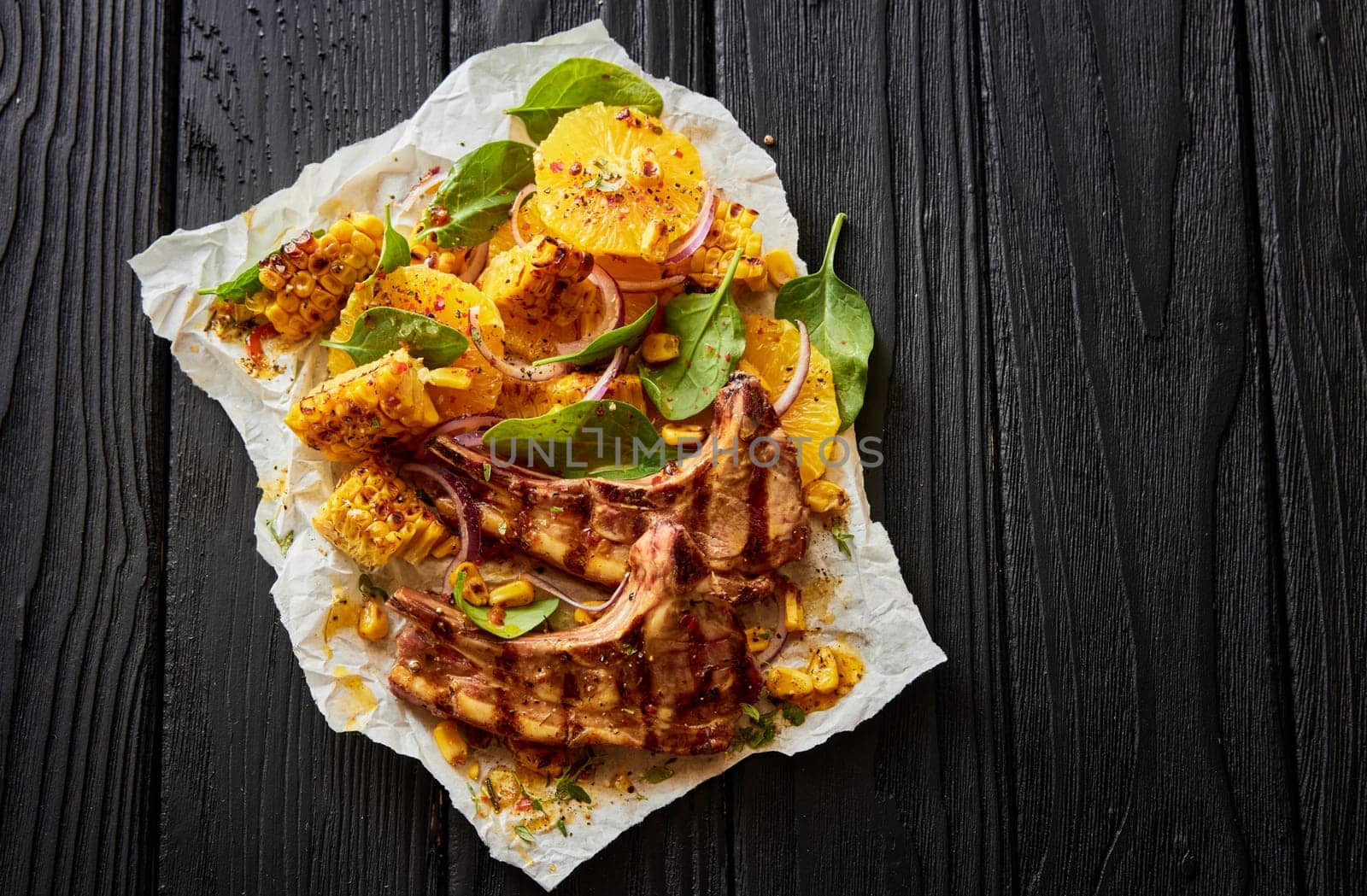 Top view of delectable grilled lamb chops with corn slices and green spinach leaves served on white parchment on black wooden background