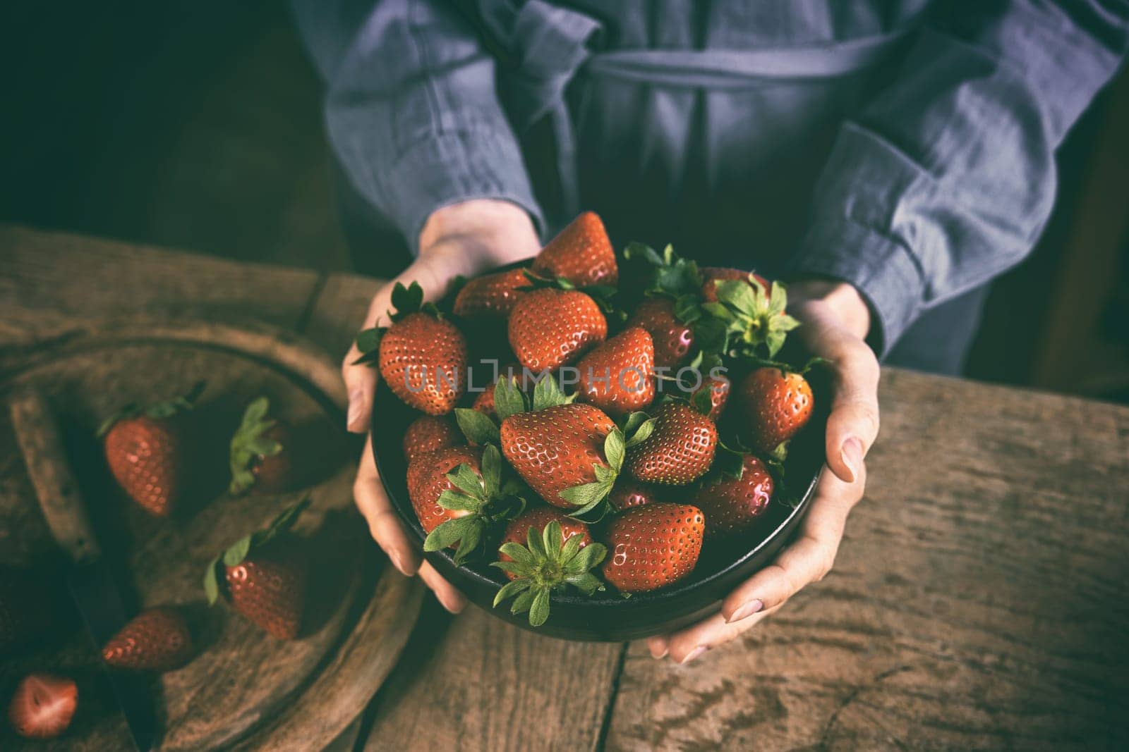 High angle of crop anonymous female in casual dress showing bowl of delicious fresh ripe strawberries standing at wooden table in kitchen in daytime